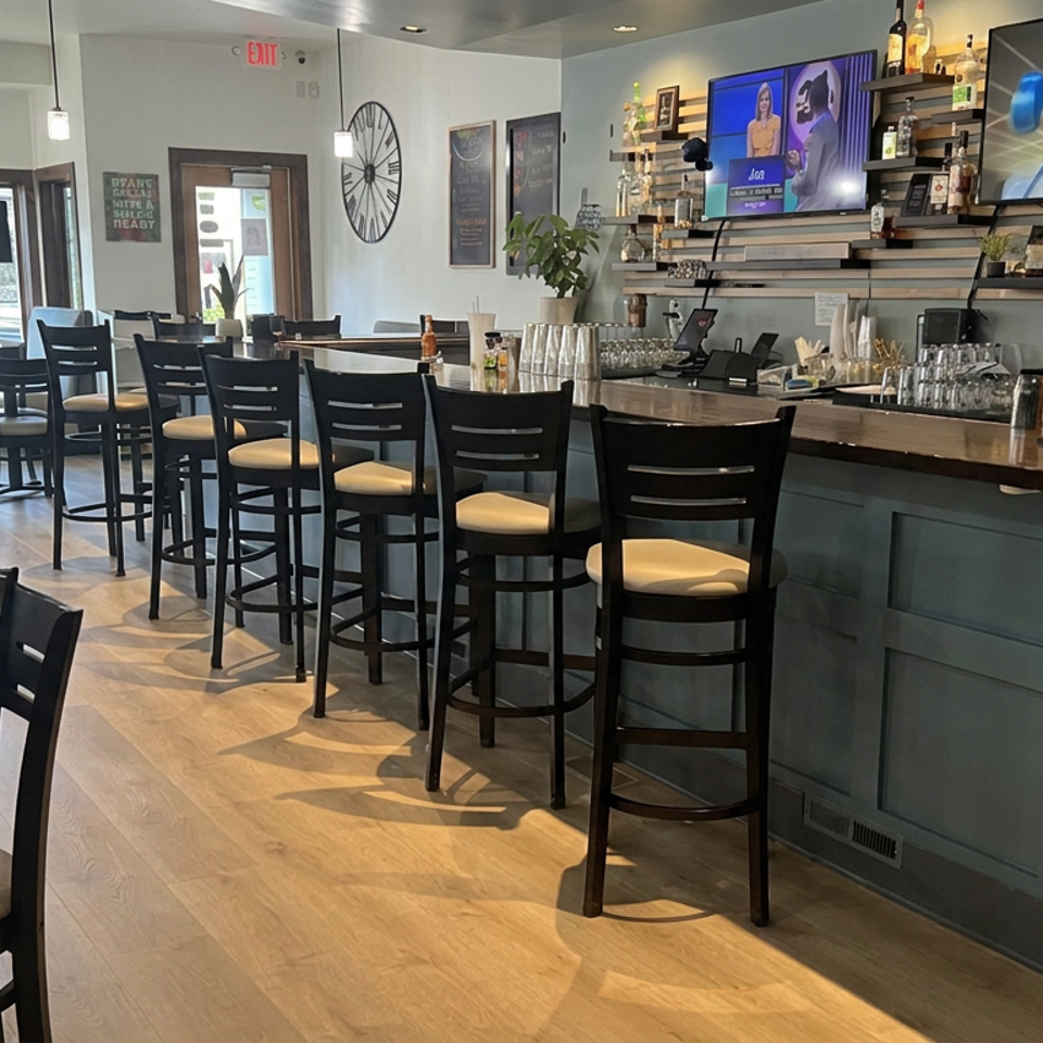 Bright restaurant dining room with black tables and chairs, a well-stocked bar, televisions, and large street-facing windows.
